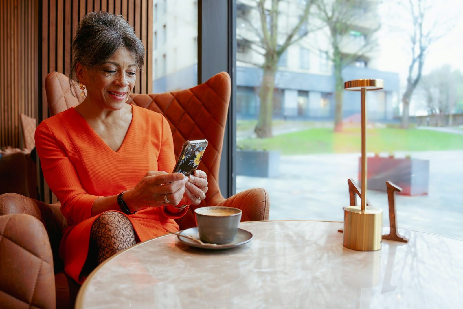 Een vrouw in een oranje jurk zit in een leren stoel bij het raam van een koffiezaak en typt glimlachend op haar telefoon